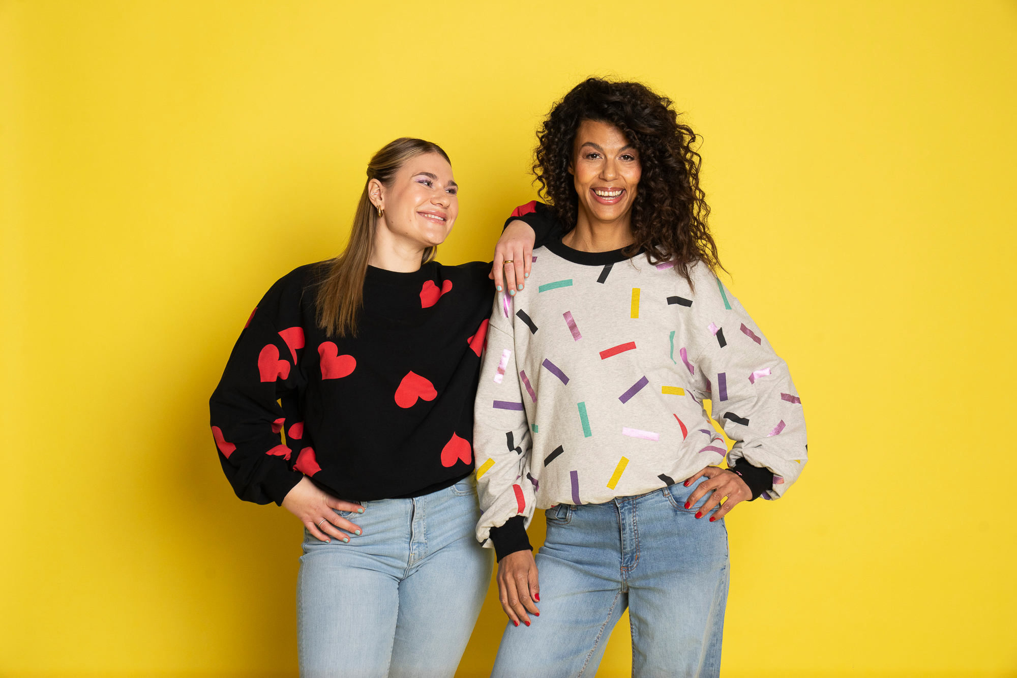 Två modeller med olika längd står framför en gul bakgrund och visar upp Celebrate Love-sweatshirts. Den kortare modellen bär en svart sweatshirt med röda hjärtan, medan den längre modellen bär en grå sweatshirt med färgglatt konfettimönster. Båda tröjorna har en avslappnad, oversized passform med vida ärmar och mudd som slutar ungefär vid höften.