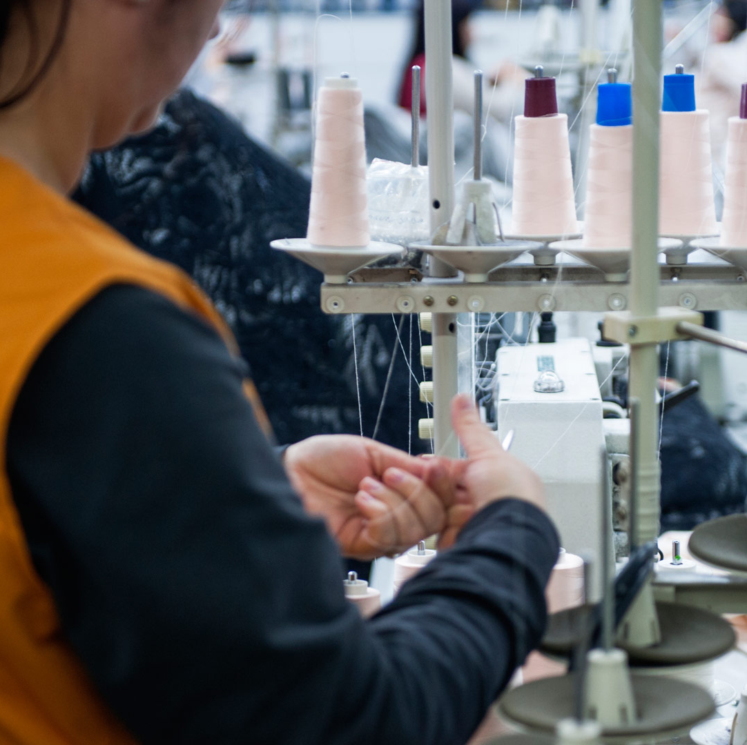 En person i orange väst arbetar vid en symaskin i en textilproduktionsmiljö. Framför maskinen syns flera trådrullar i ljusa färger och i bakgrunden pågår sömnadsarbete.