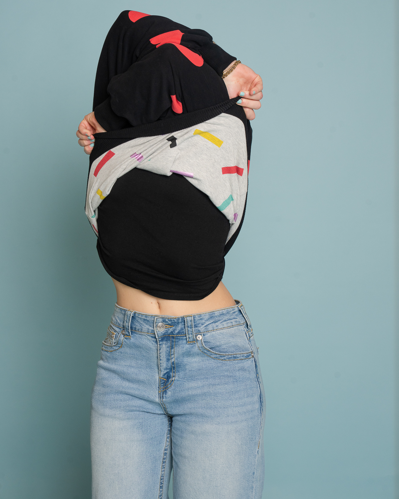 En modell i ljusa jeans drar en svart Celebrate Love-sweatshirt med röda hjärtan över huvudet. Tröjan är vänd så att det gråa fodret med färgglatt konfettimönster syns, vilket visar att plagget är vändbart. Bilden är tagen mot en ljusblå bakgrund.