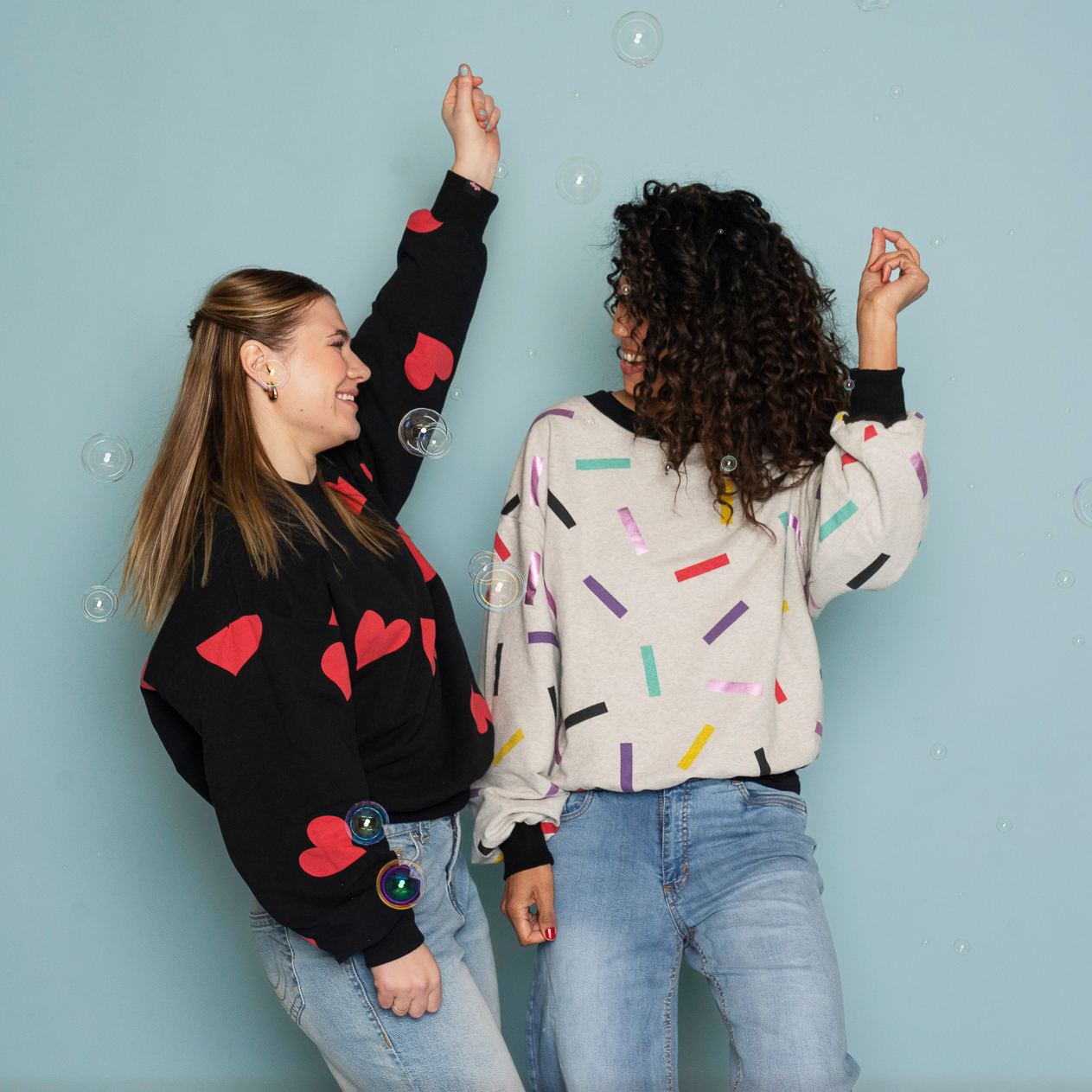 Två modeller dansar framför en ljusblå bakgrund med såpbubblor och visar upp Celebrate Love-sweatshirts. Den kortare modellen bär en svart sweatshirt med röda hjärtan, medan Vivi, som är något längre, bär en grå sweatshirt med färgglatt konfettimönster. Båda tröjorna har en avslappnad, oversized passform med vida ärmar och mudd vid höften.