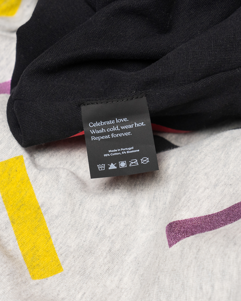 Närbild på en grå Celebrate Love-sweatshirt med färgglatt konfettimönster i gult, metalliskt lila, och svart. Bilden visar tygets struktur, den svarta mudden och plaggets etikett med texten ”Celebrate love. Wash cold, wear hot. Repeat forever.”