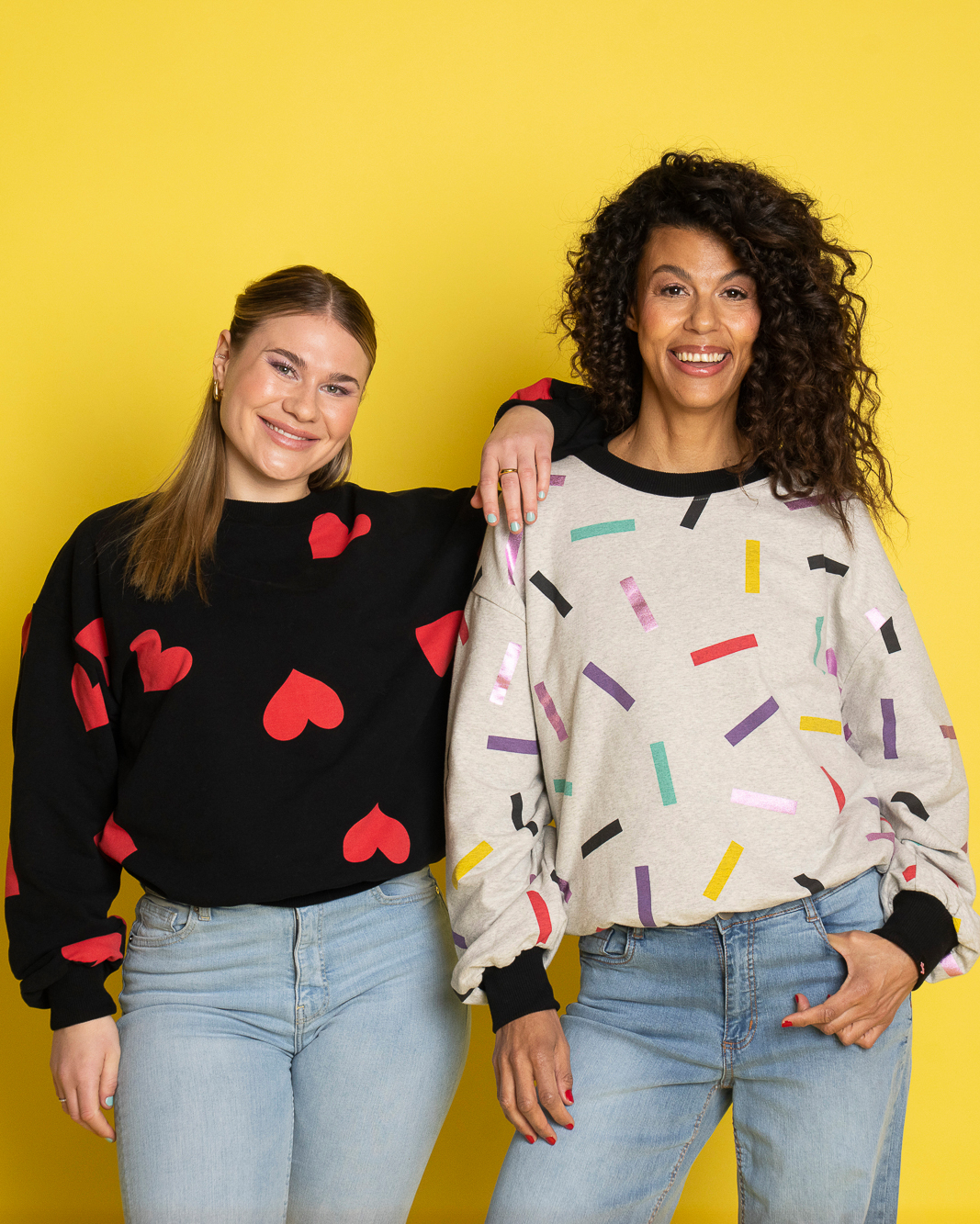 Två modeller med olika längd visar upp sweatshirts framför en gul bakgrund. Den kortare modellen bär en svart Celebrate Love-sweatshirt med röda hjärtan, medan Vivi, som är något längre, bär en grå Celebrate Love-sweatshirt med färgglatt konfettimönster. Båda tröjorna har en avslappnad, oversized passform med vida ärmar och mudd som slutar ungefär vid höften.