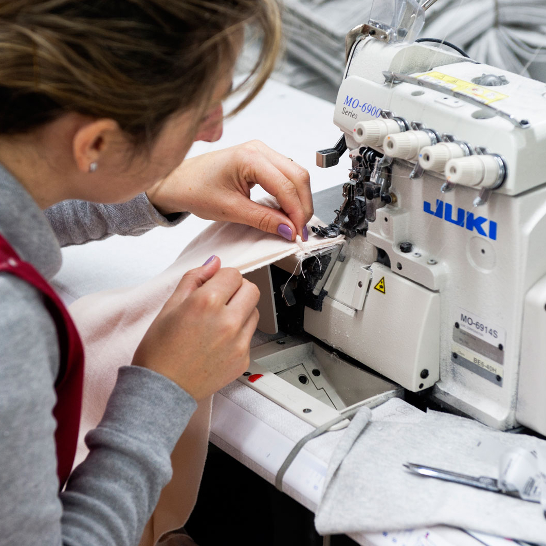 En person i röd väst arbetar vid en industrisymaskin och syr i ett ljust tyg. På arbetsbordet ligger textilier, sax och sytråd i en produktionsmiljö.