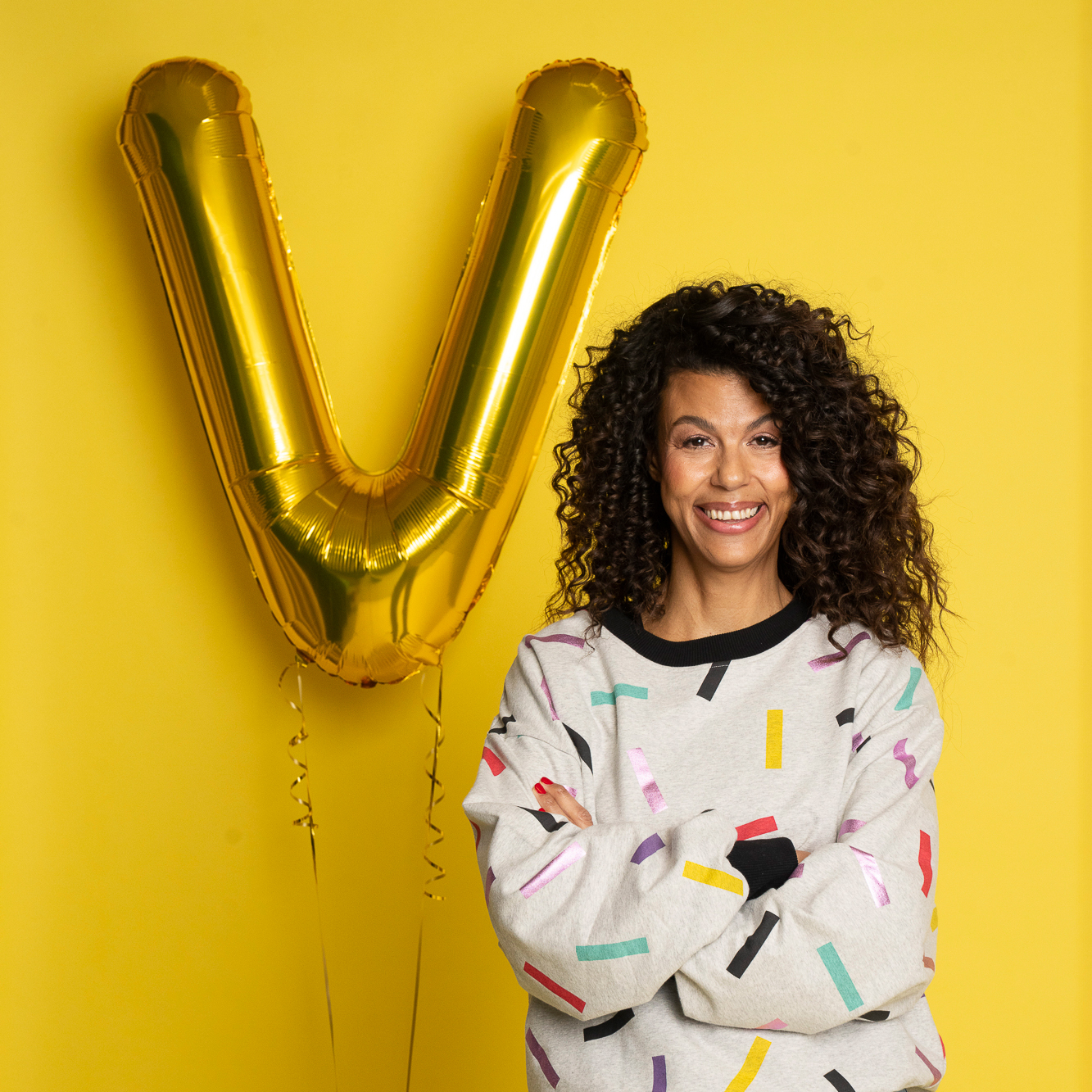 Vivi står framför en gul bakgrund bredvid en guldfärgad ballong formad som bokstaven V. Hon har armarna i kors och bär en grå sweatshirt med färgglatt konfettimönster och ljusa jeans.